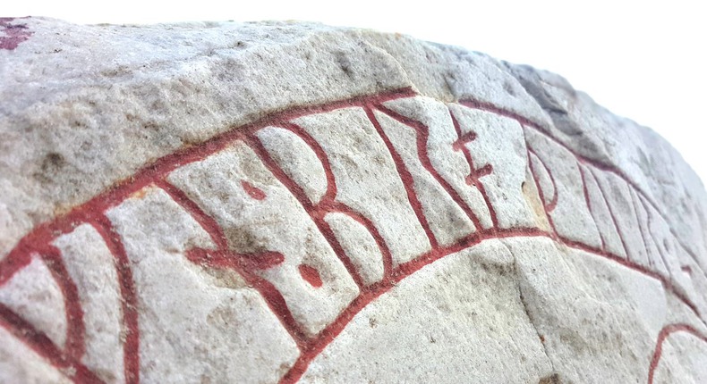 Övre delen av en runsten. Runorna är skrivna i rött i en båge som följer stenens rundade form.