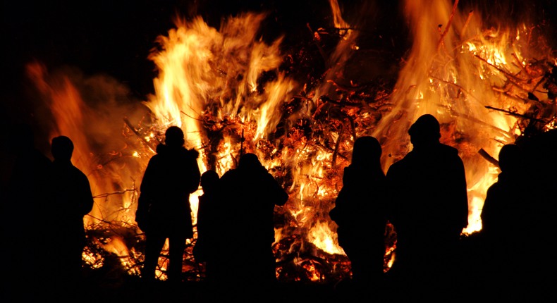 En stor brasa brinner om kvällen. Framför den syns siluetter av personer som tittar på elden.